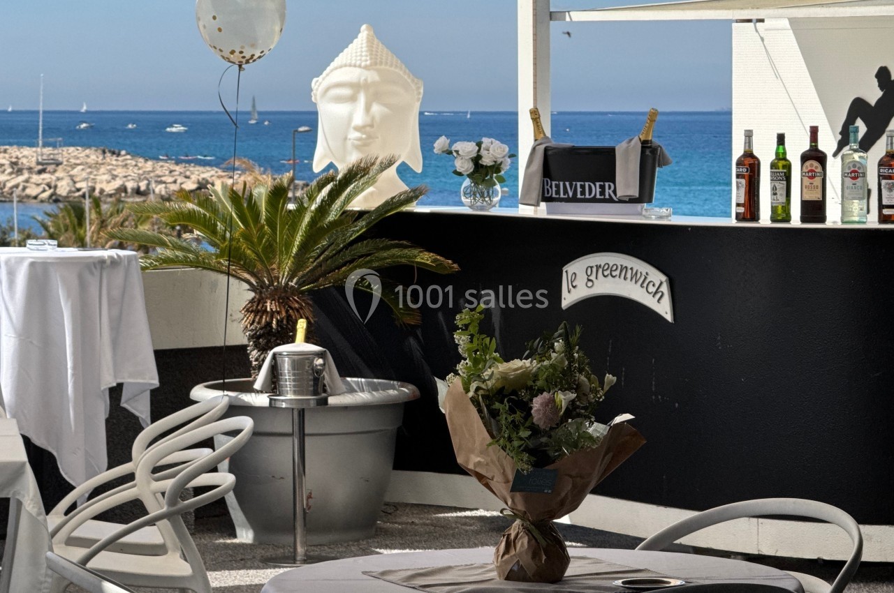 Bar extérieur avec vue sur la mer, décoré de plantes, statues et bouteilles, table dressée au premier plan.