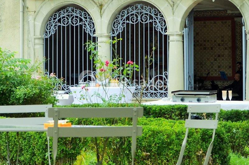 Chaises blanches et table dans un jardin, devant des arches avec grilles en fer forgé et un mur décoré de mosaïques.