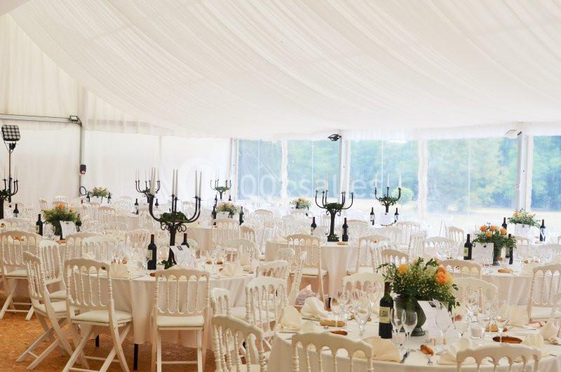 Salle de réception décorée avec des tables rondes, nappes blanches, chandeliers noirs et vue sur un extérieur verdoyant.