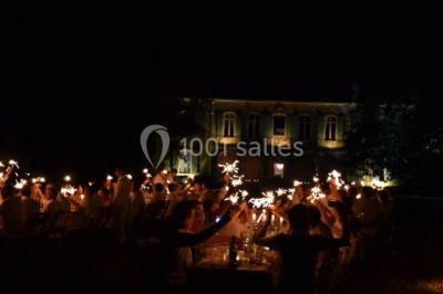 Des personnes en tenue blanche brandissent des cierges magiques lors d'un dîner en plein air devant un bâtiment éclairé.