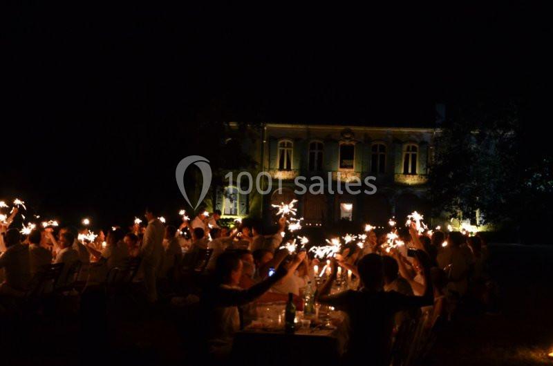 Des personnes en tenue blanche brandissent des cierges magiques lors d'un dîner en plein air devant un bâtiment éclairé.