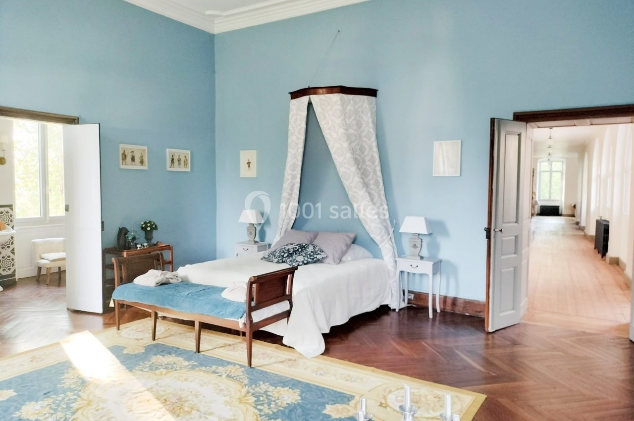 Chambre lumineuse avec murs bleus, lit à baldaquin, parquet en bois et tapis décoratif, portes ouvertes sur un couloir.
