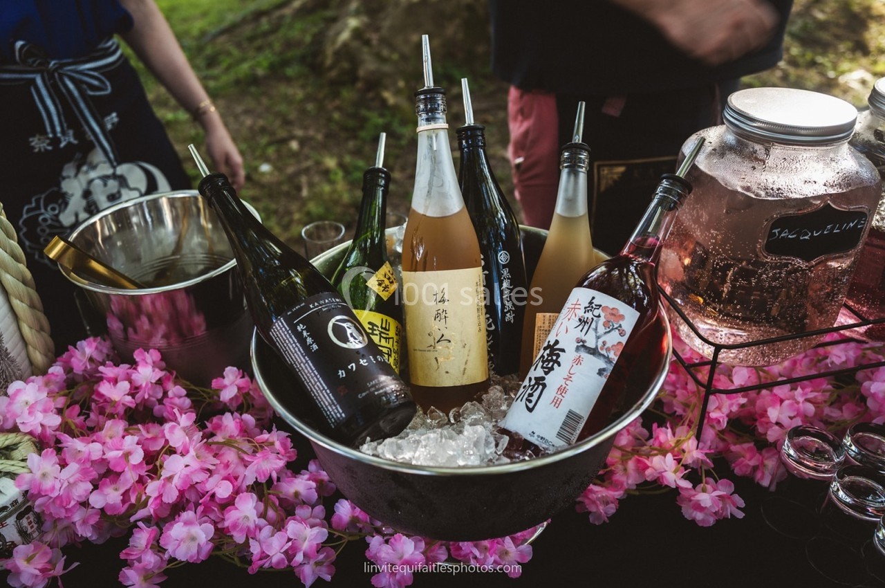 Bouteille de saké et autres boissons disposées sur de la glace dans un seau, entourées de fleurs roses décoratives.
