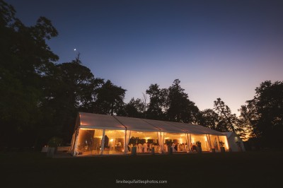 Vue d'une tente éclairée de nuit dans un jardin, entourée d'arbres sous un ciel crépusculaire.