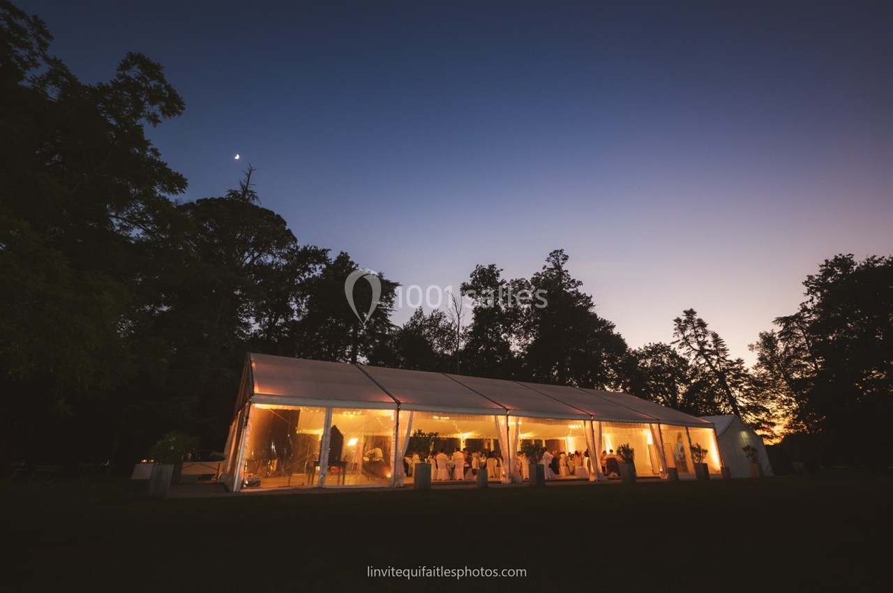 Vue d'une tente éclairée de nuit dans un jardin, entourée d'arbres sous un ciel crépusculaire.