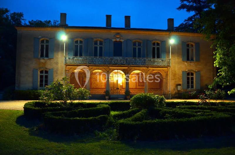 Façade d'un bâtiment ancien éclairé la nuit, avec un jardin à haies taillées au premier plan.