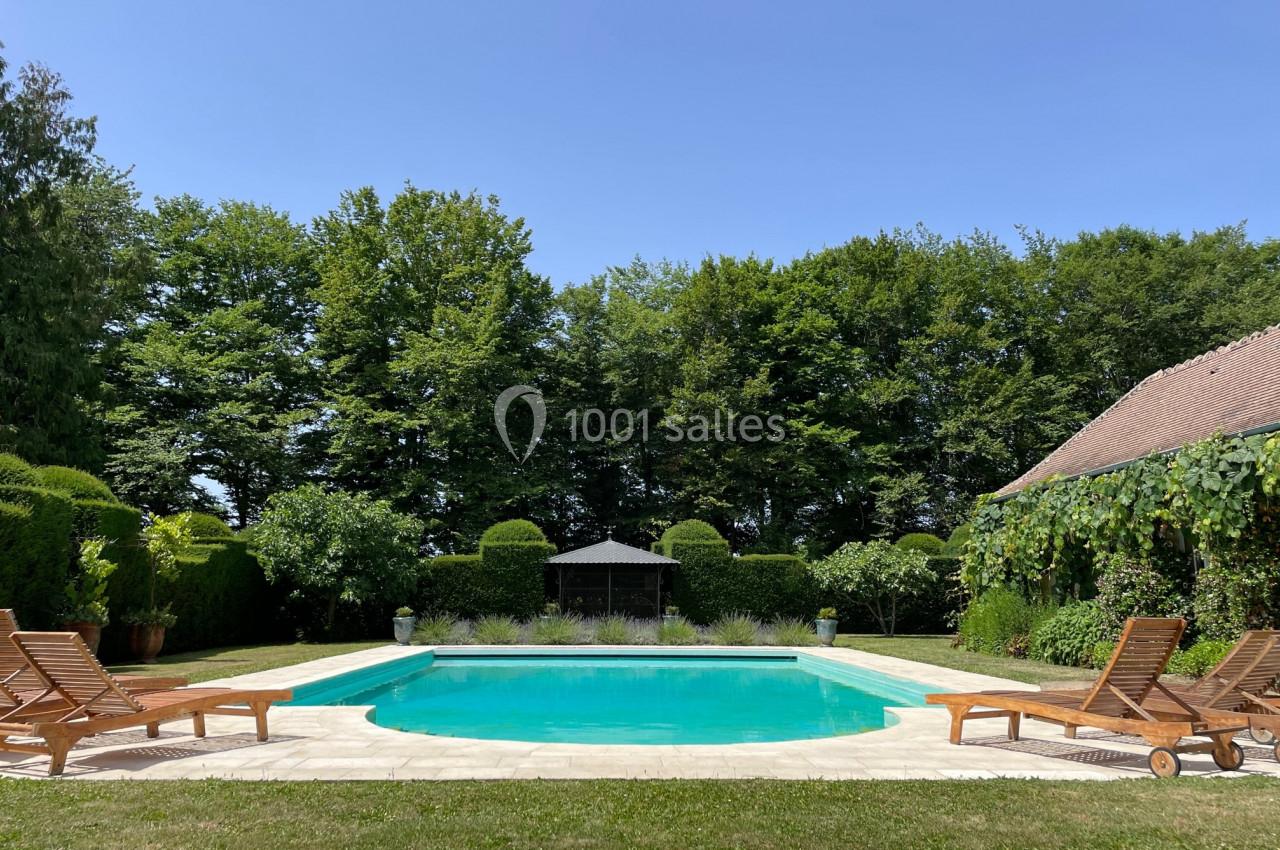 Piscine extérieure entourée de chaises longues en bois, pelouse, arbres et cabanon sous un ciel dégagé.