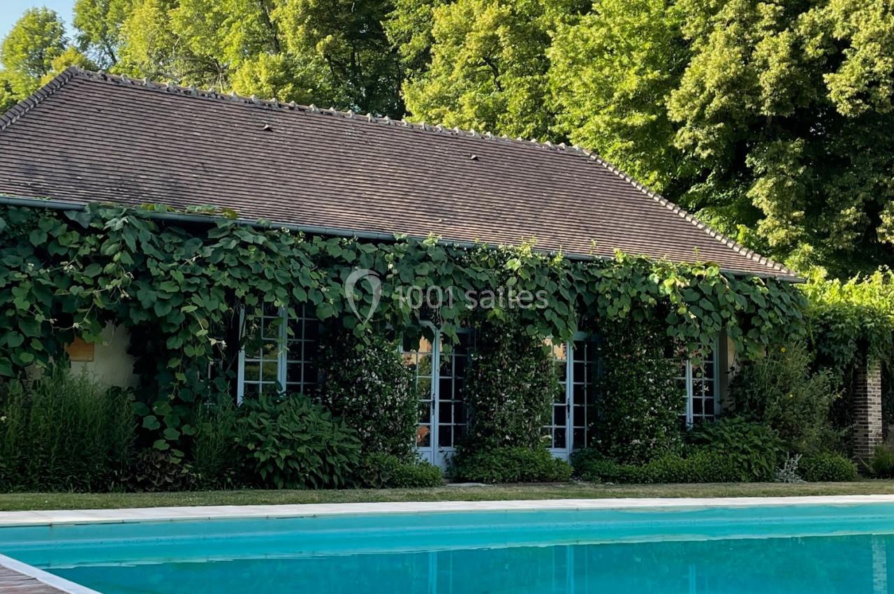 Maison entourée de verdure avec une piscine au premier plan et des arbres en arrière-plan sous un ciel dégagé.