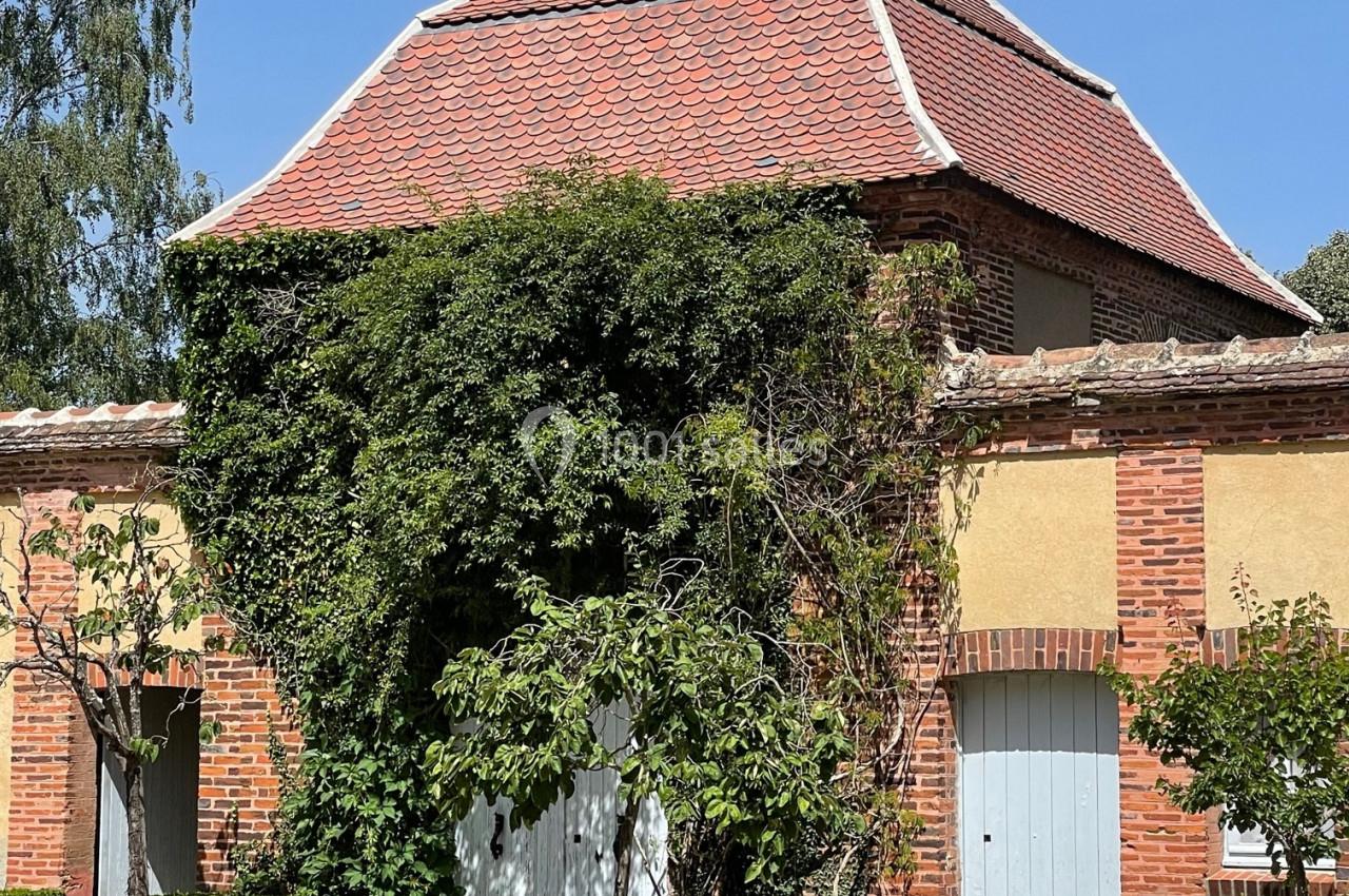 Bâtiment en briques avec toit en tuiles rouges, entouré de plantes grimpantes et d'arbustes taillés.