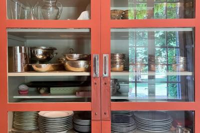 Miniature Location salle Triguères (Loiret) - Manoir du Grand Courtoiseau et ses Jardins Remarquables #37 Vaisselier rouge avec portes vitrées, contenant des verres, plats, bols et assiettes en céramique et en métal.