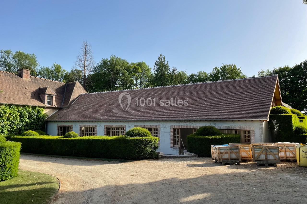 Bâtiment avec toit en tuiles entouré de haies taillées, sur une allée en gravier, par temps ensoleillé.