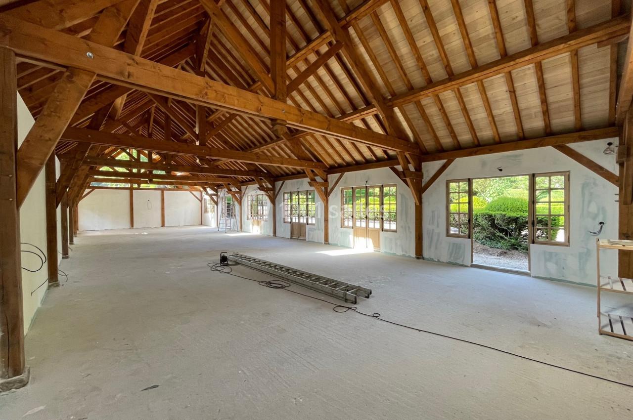 Intérieur d'un grand bâtiment en rénovation avec charpente en bois apparente et fenêtres donnant sur un jardin.