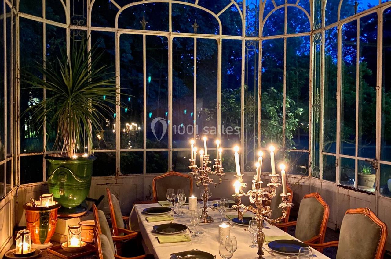 Table dressée avec chandeliers allumés dans une verrière élégante, entourée de végétation visible à travers les vitres.