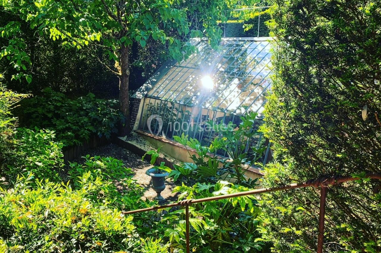 Serre vitrée entourée de végétation luxuriante sous une lumière ensoleillée.