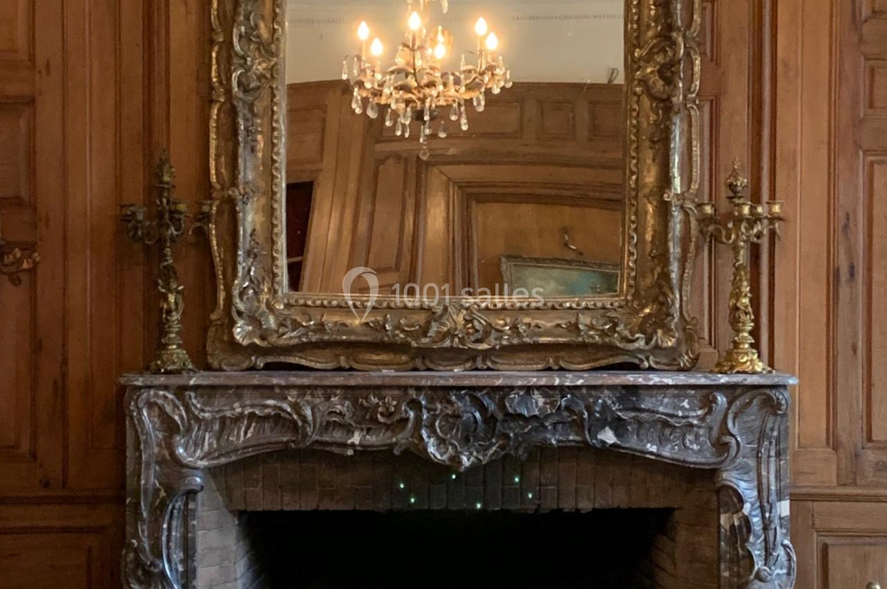 Miroir doré au-dessus d'une cheminée en marbre sculpté, entouré de boiseries et éclairé par un lustre.