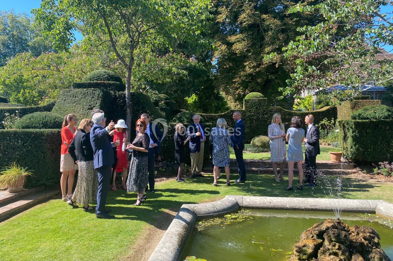 Groupe de personnes discutant dans un jardin avec une fontaine et des haies taillées en arrière-plan.