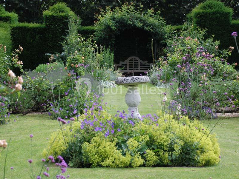 Jardin fleuri avec une fontaine centrale en pierre, entourée de buissons taillés et de fleurs colorées.