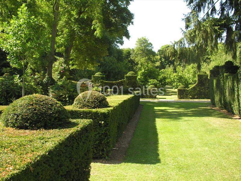 Jardin avec haies taillées, arbustes en topiaire et pelouse bordée d'arbres sous un ciel dégagé.