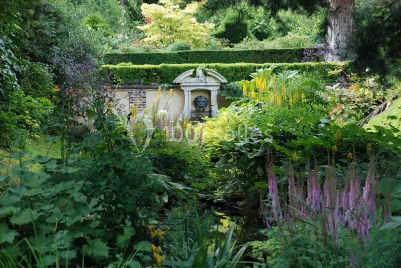 Vue d'un jardin verdoyant avec une fontaine ornée d'un masque en pierre, entourée de plantes et de fleurs.