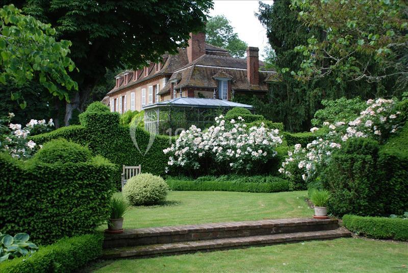 Jardin verdoyant avec haies taillées, rosiers en fleurs et une maison en briques rouges en arrière-plan.