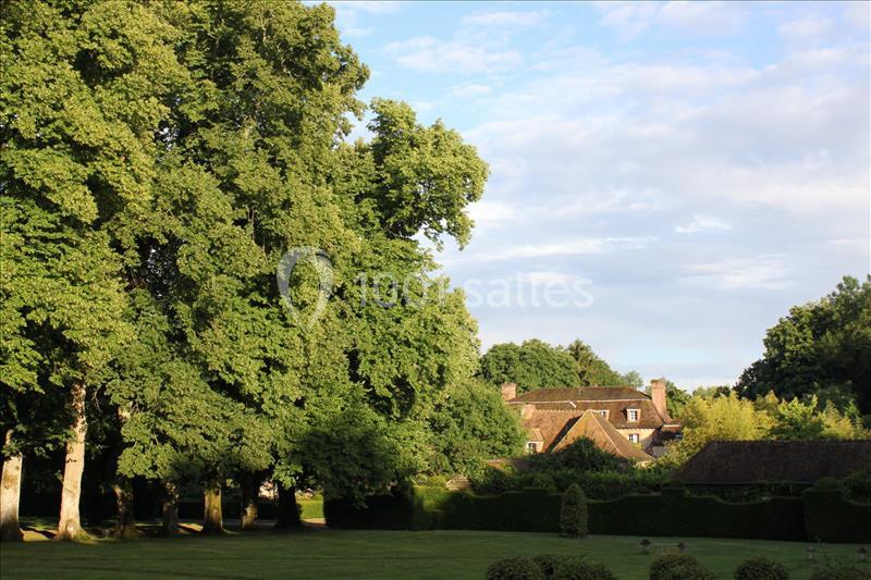 Vue d'une maison entourée d'arbres et de haies dans un jardin verdoyant sous un ciel partiellement dégagé.