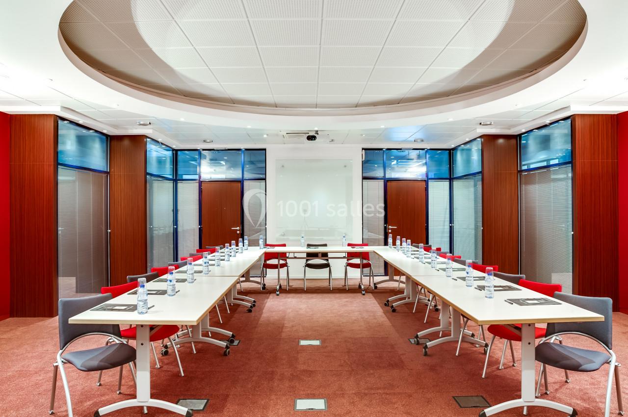 Salle de réunion moderne avec tables disposées en U, chaises rouges et grises, bouteilles d'eau et éclairage lumineux.