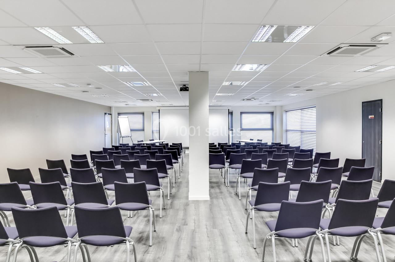 Salle de conférence moderne avec rangées de chaises vides, éclairée par des fenêtres et des lumières au plafond.