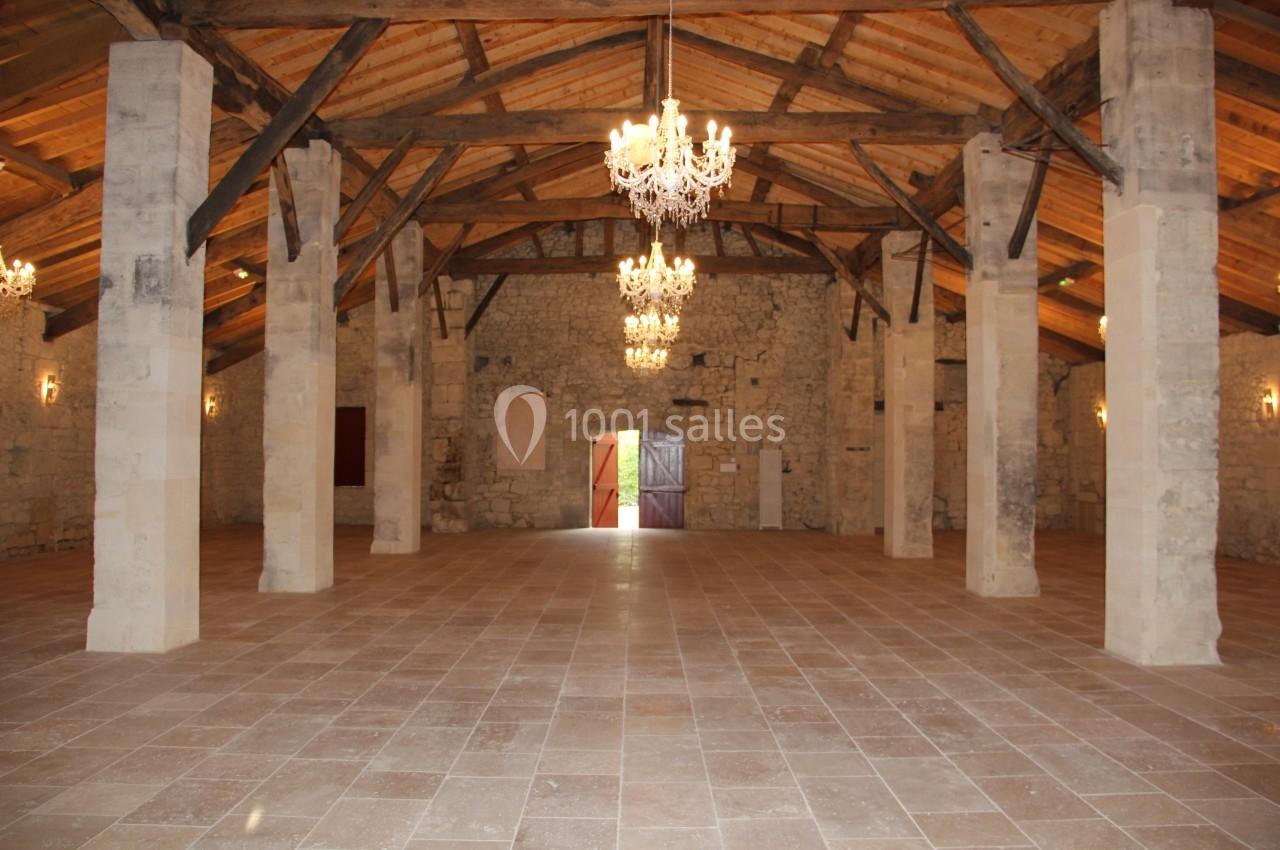 Salle spacieuse avec murs en pierre, poutres en bois apparentes et lustres suspendus, éclairée par une porte ouverte.