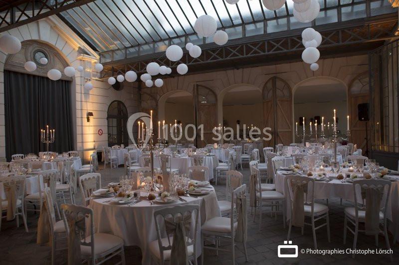 Salle de réception élégante avec tables dressées, chandeliers et lanternes suspendues sous une verrière.