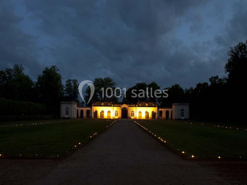 Façade d'un bâtiment illuminé la nuit, entouré d'arbres et d'une allée bordée de petites lumières au sol.