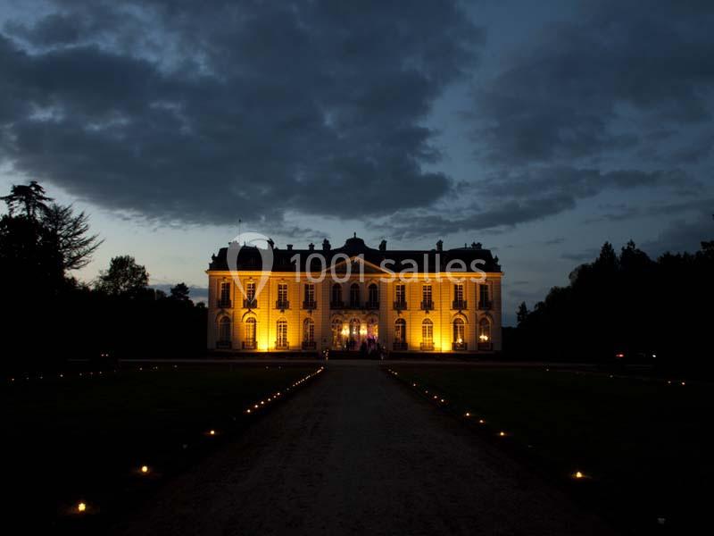 Façade d'un château illuminé la nuit, entouré de jardins sombres et d'un chemin bordé de petites lumières.