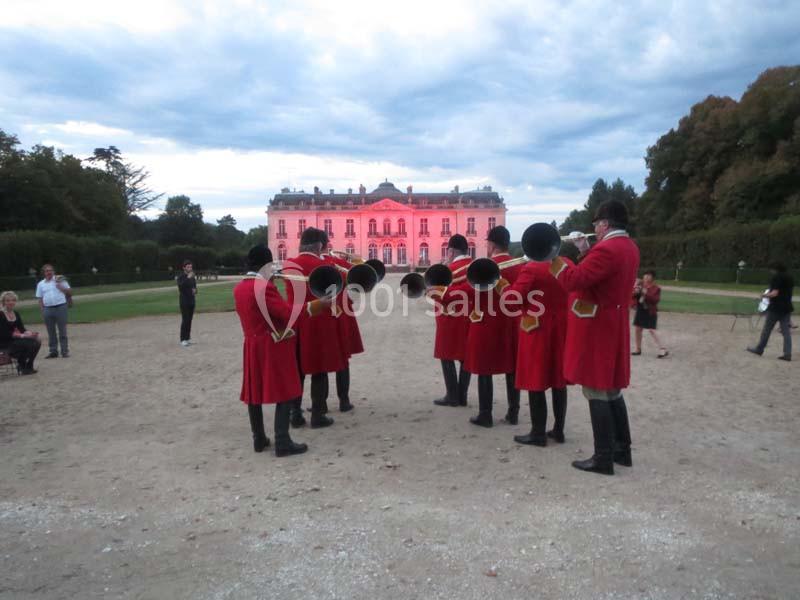 Un groupe de musiciens en tenue rouge joue du cor devant un château éclairé en rose au crépuscule.