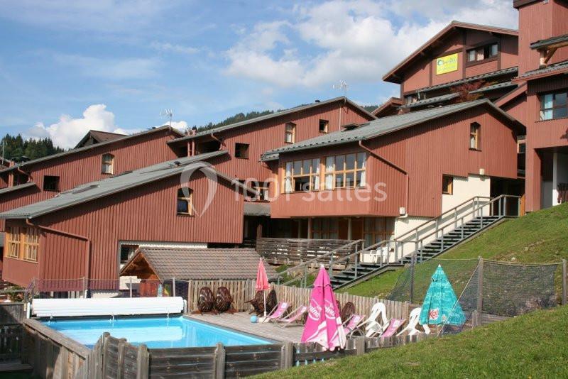 Résidence en bois avec piscine extérieure, transats et parasols colorés, située sur une colline par temps ensoleillé.