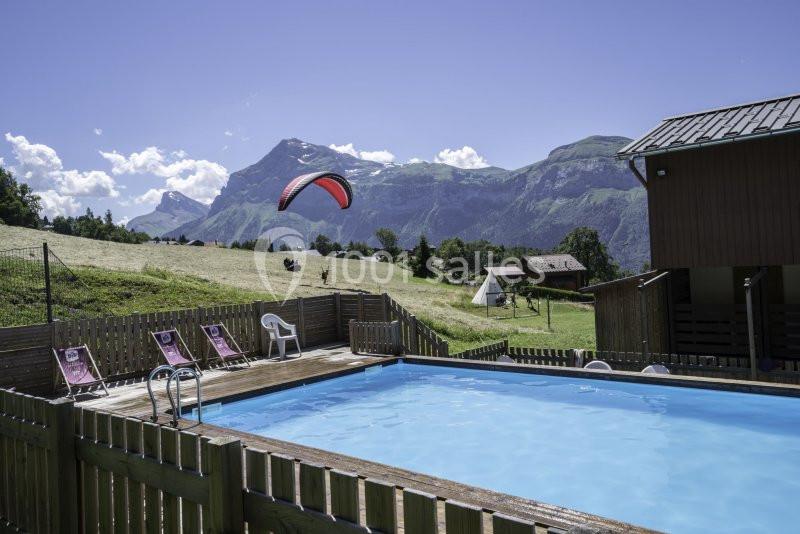 Piscine extérieure entourée d'une clôture en bois, avec vue sur des montagnes et un parapente dans le ciel.