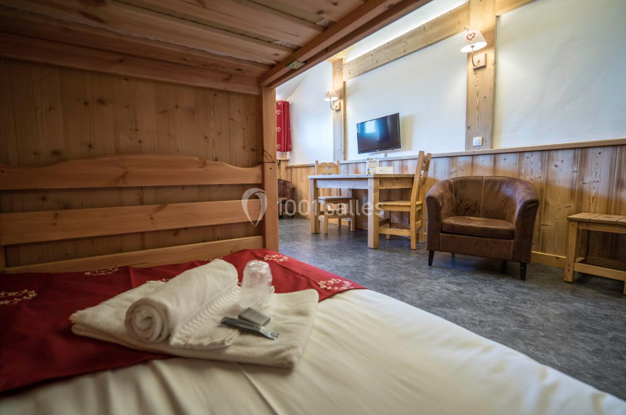 Chambre en bois avec lit, table, chaises, fauteuils, télévision murale et lumière naturelle.