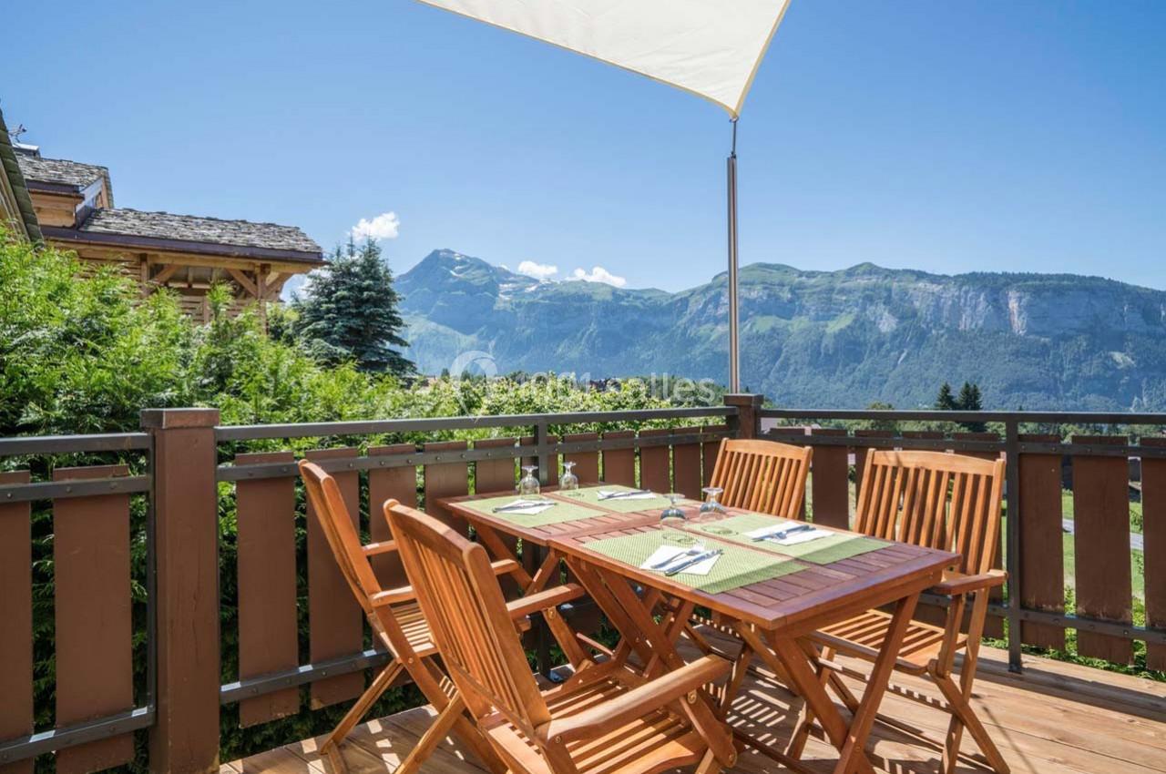 Terrasse en bois avec table et chaises, vue sur des montagnes verdoyantes sous un ciel bleu dégagé.