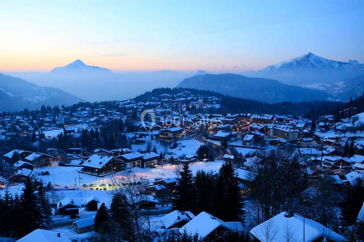 Vue d'un village de montagne enneigé au crépuscule, entouré de forêts et de sommets éclairés par une lumière douce.