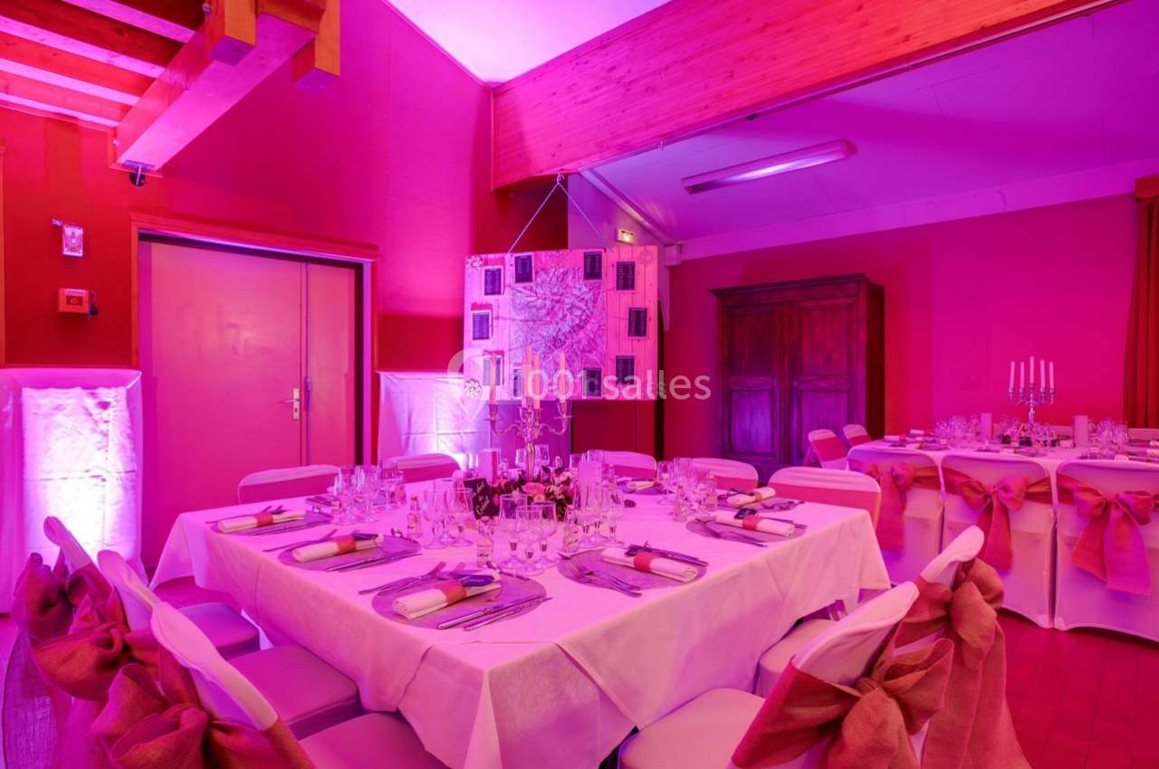 Salle de réception décorée en rose avec des tables dressées, nappes blanches et chaises ornées de nœuds roses.