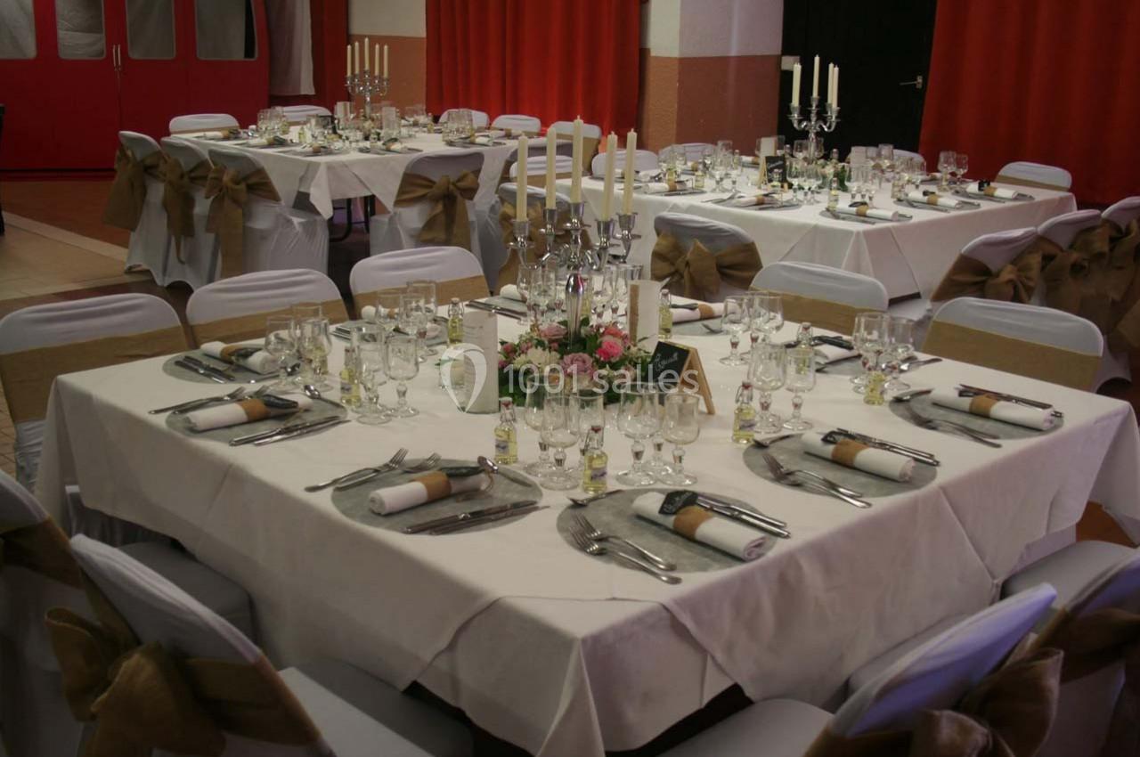 Tables élégamment dressées avec nappes blanches, bougies, fleurs et chaises décorées pour un événement festif en intérieur.