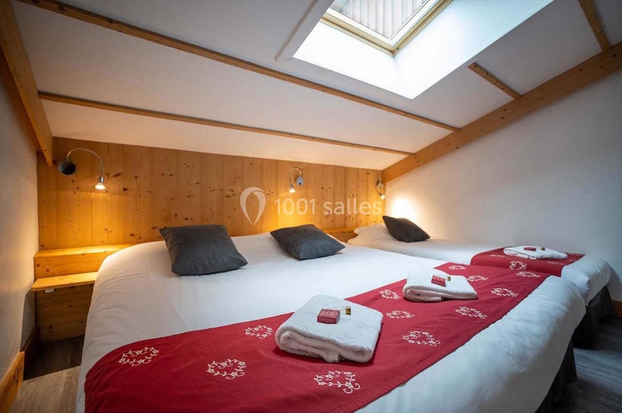 Chambre mansardée avec deux lits simples rapprochés, éclairage mural, linge plié et fenêtre de toit.