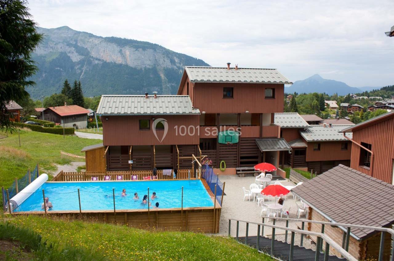 Piscine extérieure avec baigneurs, entourée de bâtiments en bois et de montagnes verdoyantes en arrière-plan.