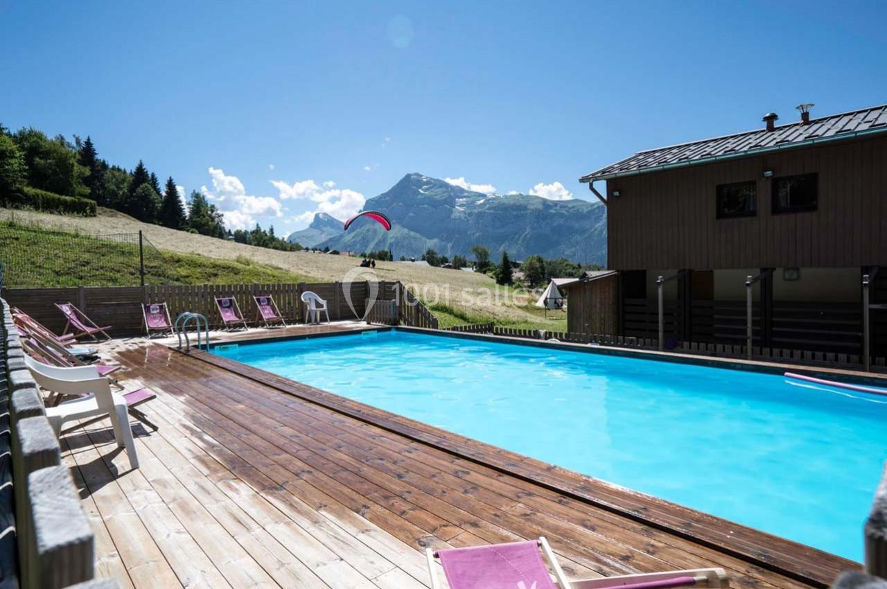 Piscine extérieure avec terrasse en bois, vue sur les montagnes et un parapente dans le ciel en arrière-plan.
