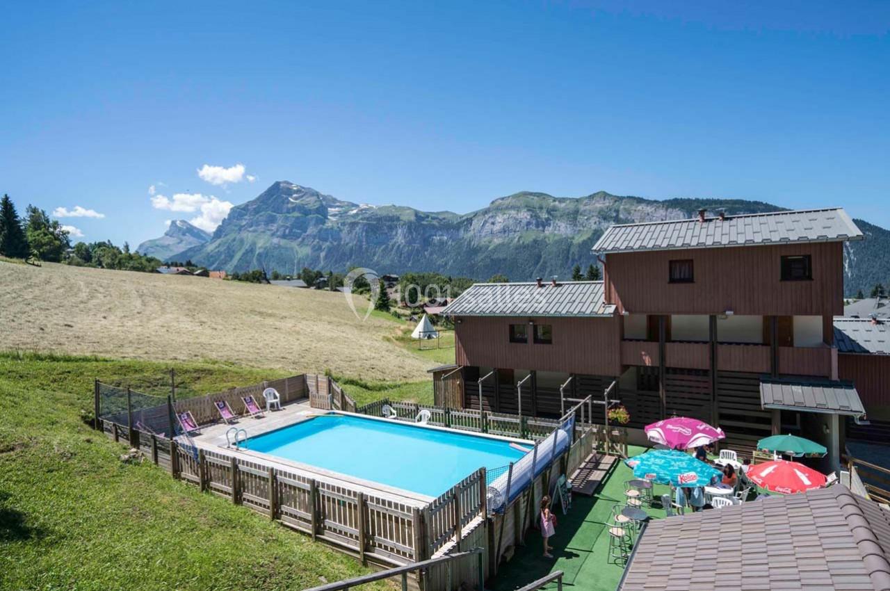 Piscine extérieure entourée de transats, située près de bâtiments en bois avec vue sur des montagnes verdoyantes.