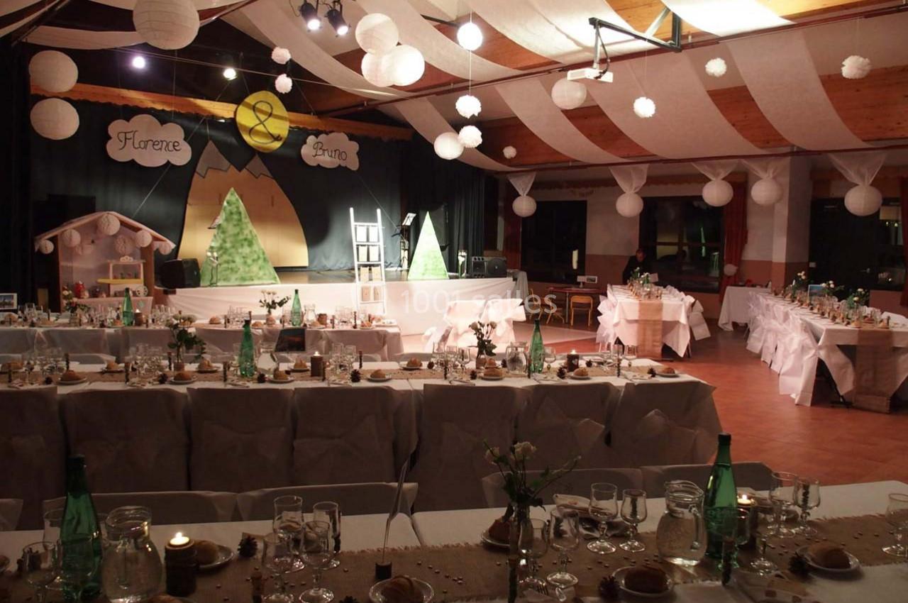 Salle décorée pour une réception avec tables dressées, guirlandes lumineuses et scène ornée de panneaux personnalisés.