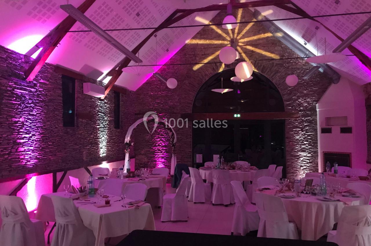 Salle de réception décorée avec des lumières roses, des tables dressées et une arche ornée de fleurs blanches.