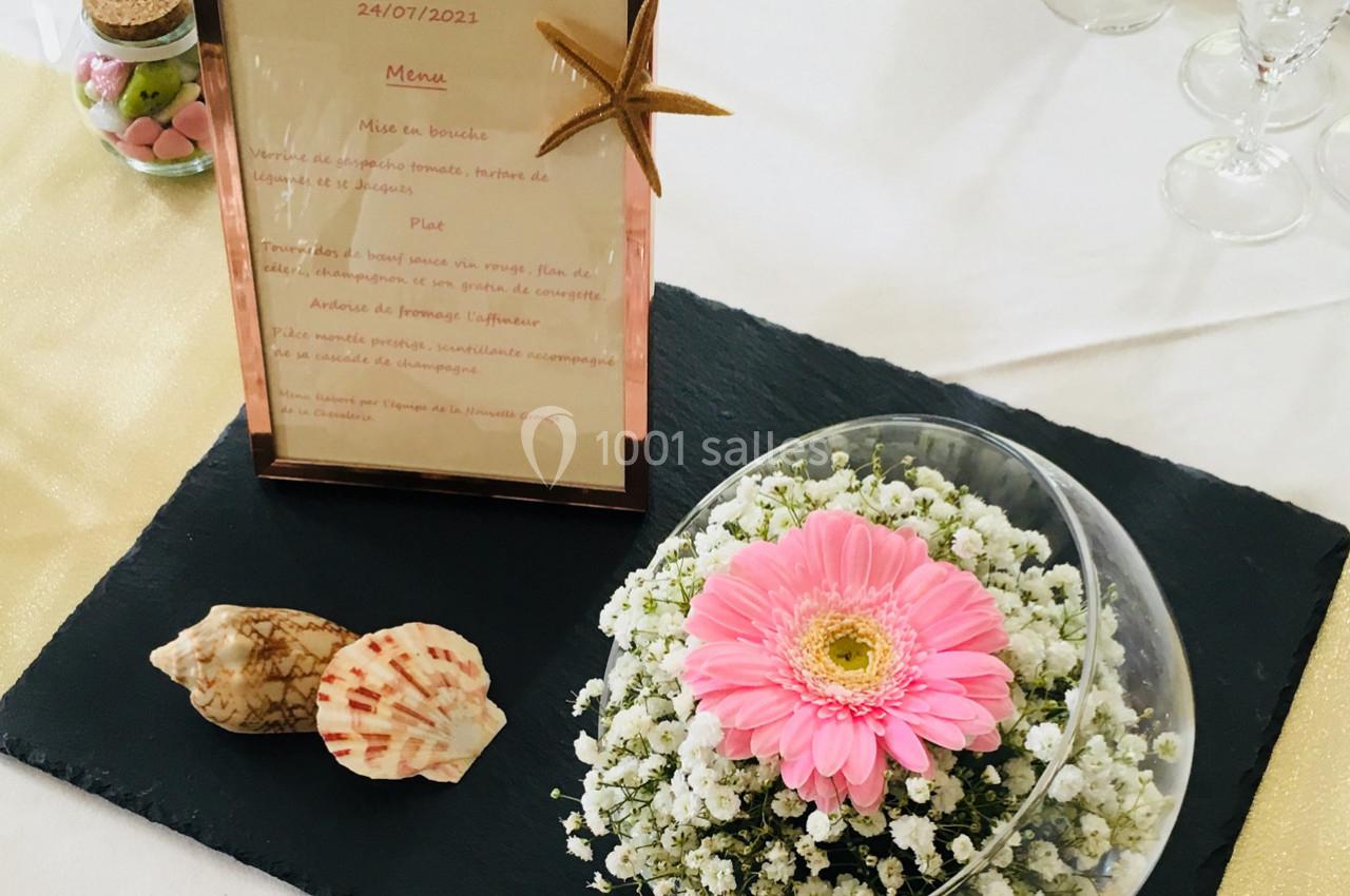 Menu encadré posé sur une table avec une décoration florale, des coquillages et des verres en arrière-plan.