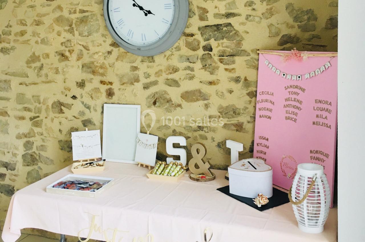 Table décorée pour un mariage avec une nappe rose, des accessoires, une urne, et un panneau de noms sur fond de mur en…