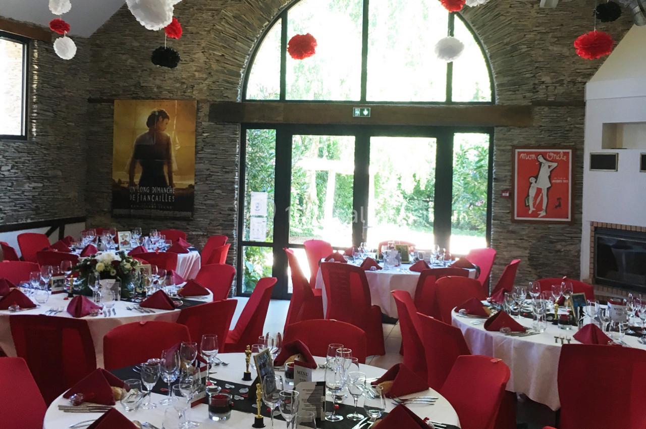 Salle de réception décorée avec des tables dressées, nappes blanches, serviettes rouges et affiches aux murs en pierre.