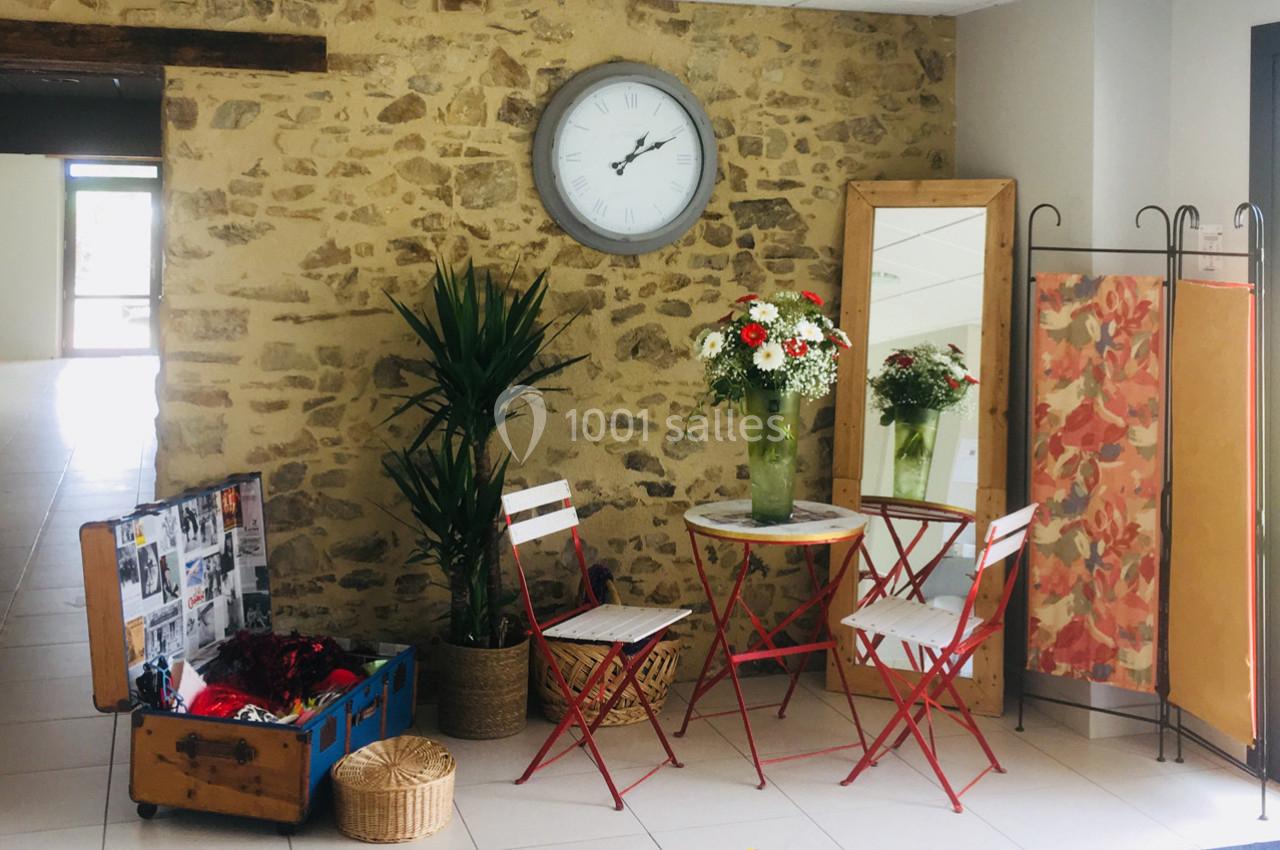 Coin décoratif avec mur en pierre, table et chaises rouges, plantes, miroir, horloge murale et coffre ouvert.