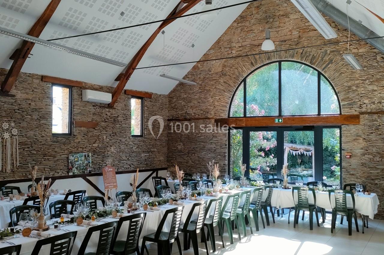 Salle de réception lumineuse avec tables décorées, poutres apparentes et grande baie vitrée donnant sur un jardin fleuri.